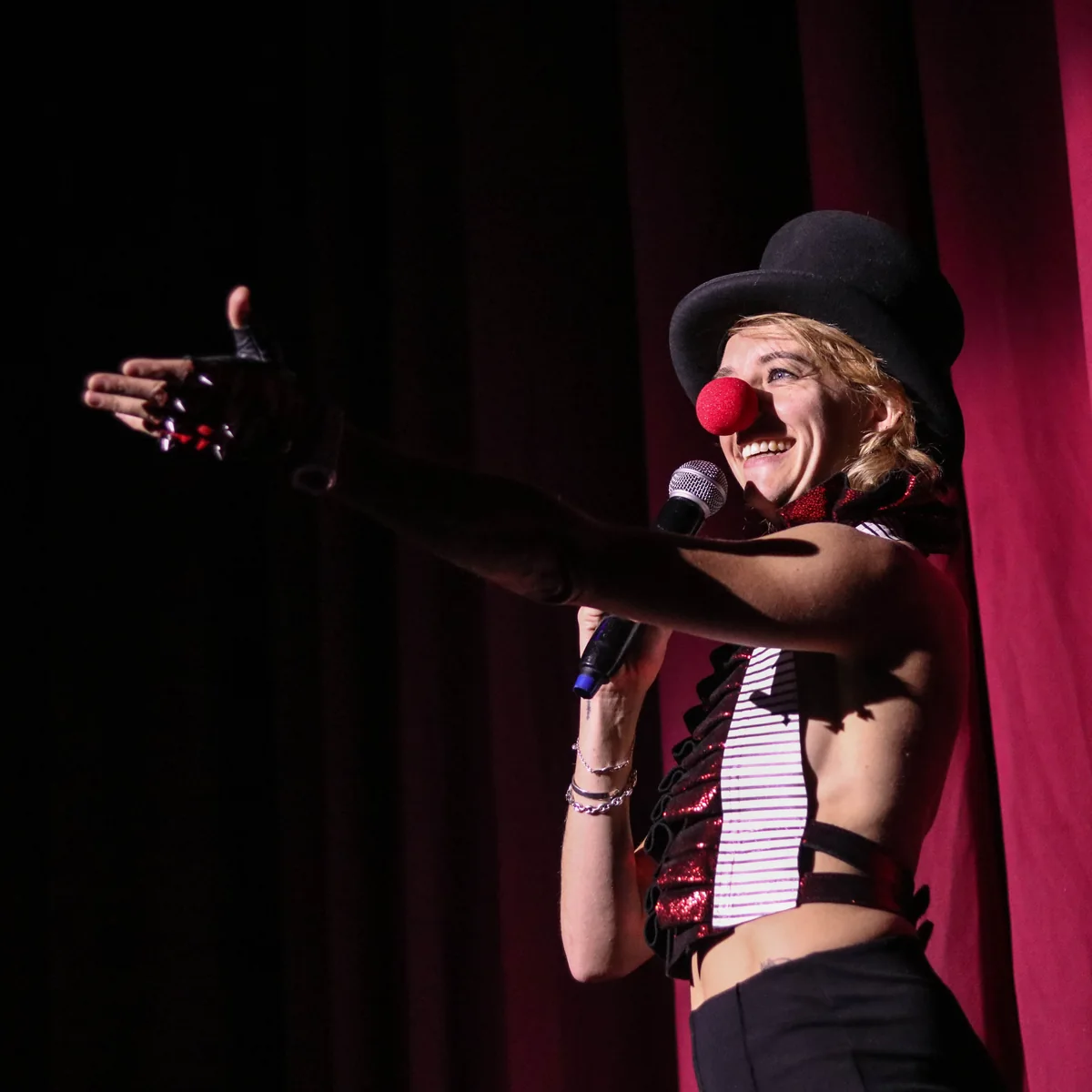 Ellie J, Clown Daddy, laughing on stage in a top hat and red nose, arm flung out, her shadow on the red curtain