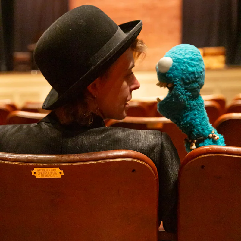 Clown Daddy in a bowler hat sitting in red theater seats beside her blue puppet Gratch