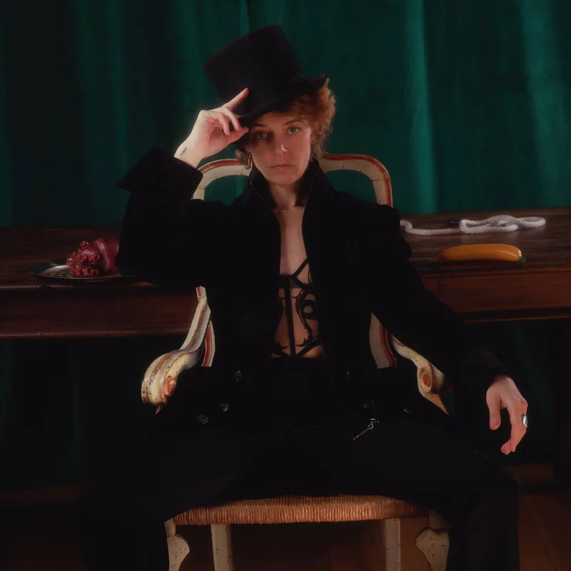Ellie J, Clown Daddy, in a black top hat and suit, seated on a wooden chair in front of a green velvet curtain