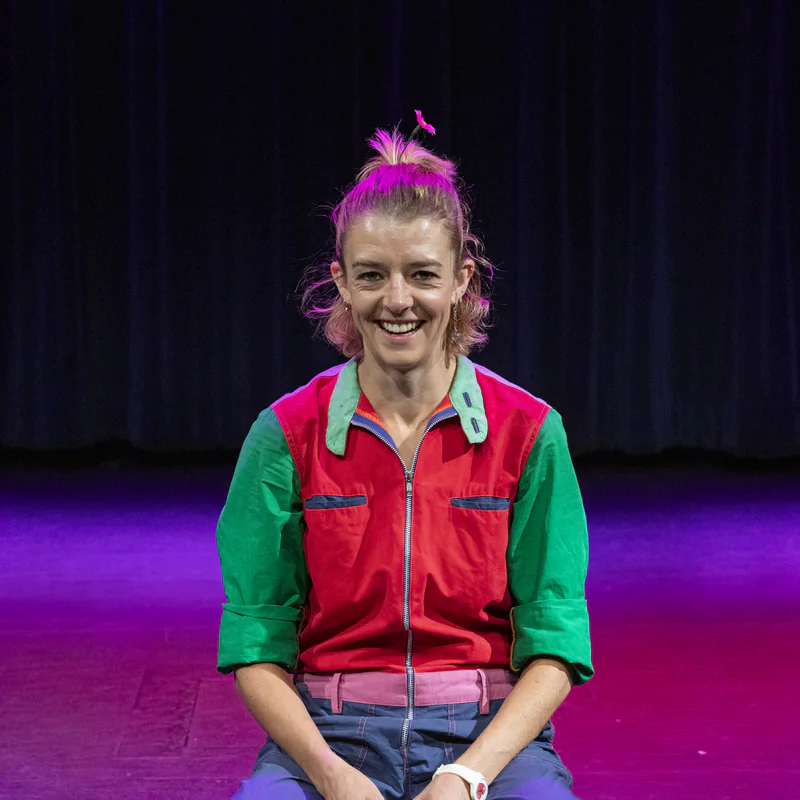 Ellie J smiling on a purple stage in a red and green jumpsuit, a red clown nose on the floor in front of her
