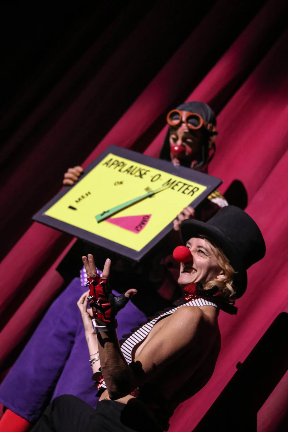 Clown Daddy hosting on stage in a top hat and red nose, holding up a framed sign in front of a red curtain